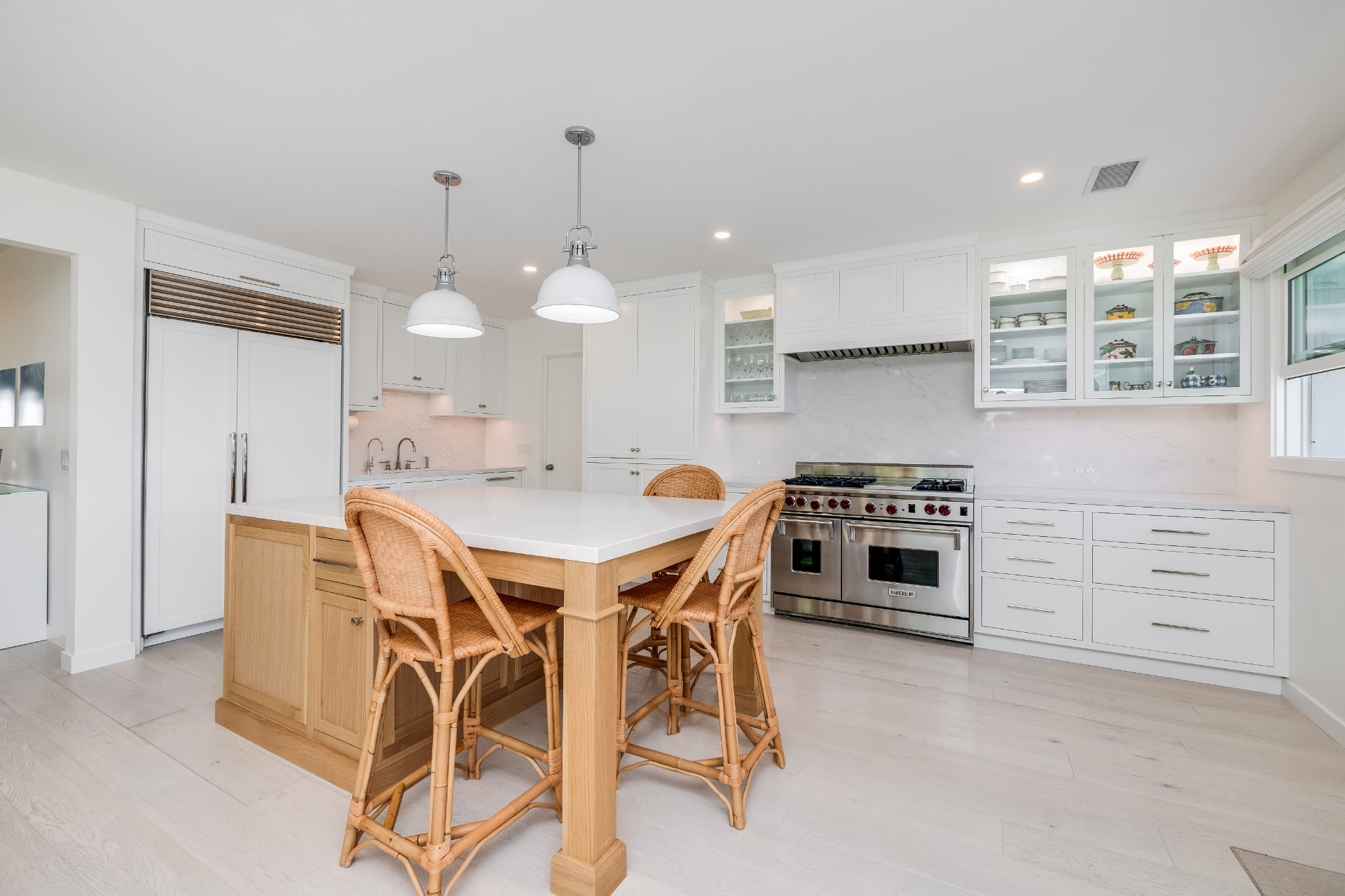 Bright white kitchen with a light wood island, woven bar stools, pendant lighting and professional stainless range